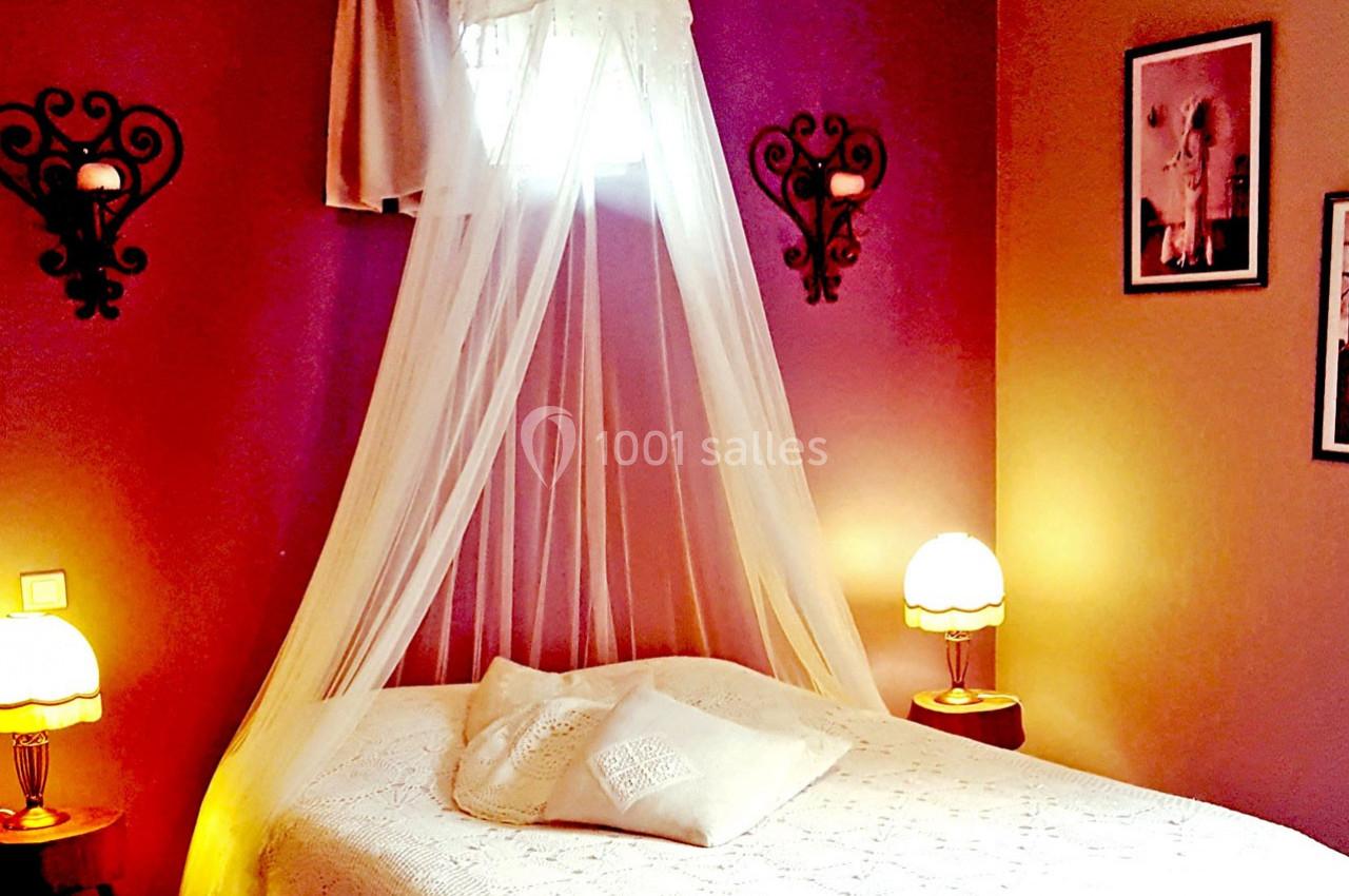 Lit à baldaquin avec voilage blanc dans une chambre aux murs violets, éclairée par des lampes de chevet.