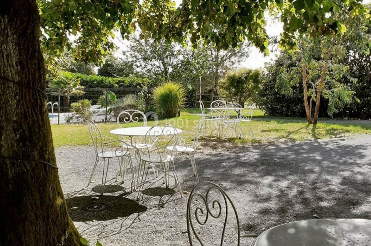 Chaises et tables blanches en fer forgé disposées sous des arbres dans un jardin ensoleillé.