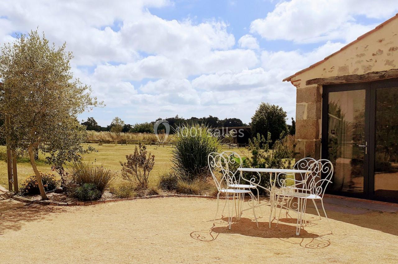 Table et chaises blanches en fer forgé sur une terrasse en gravier, avec vue sur un jardin et des champs sous un ciel…