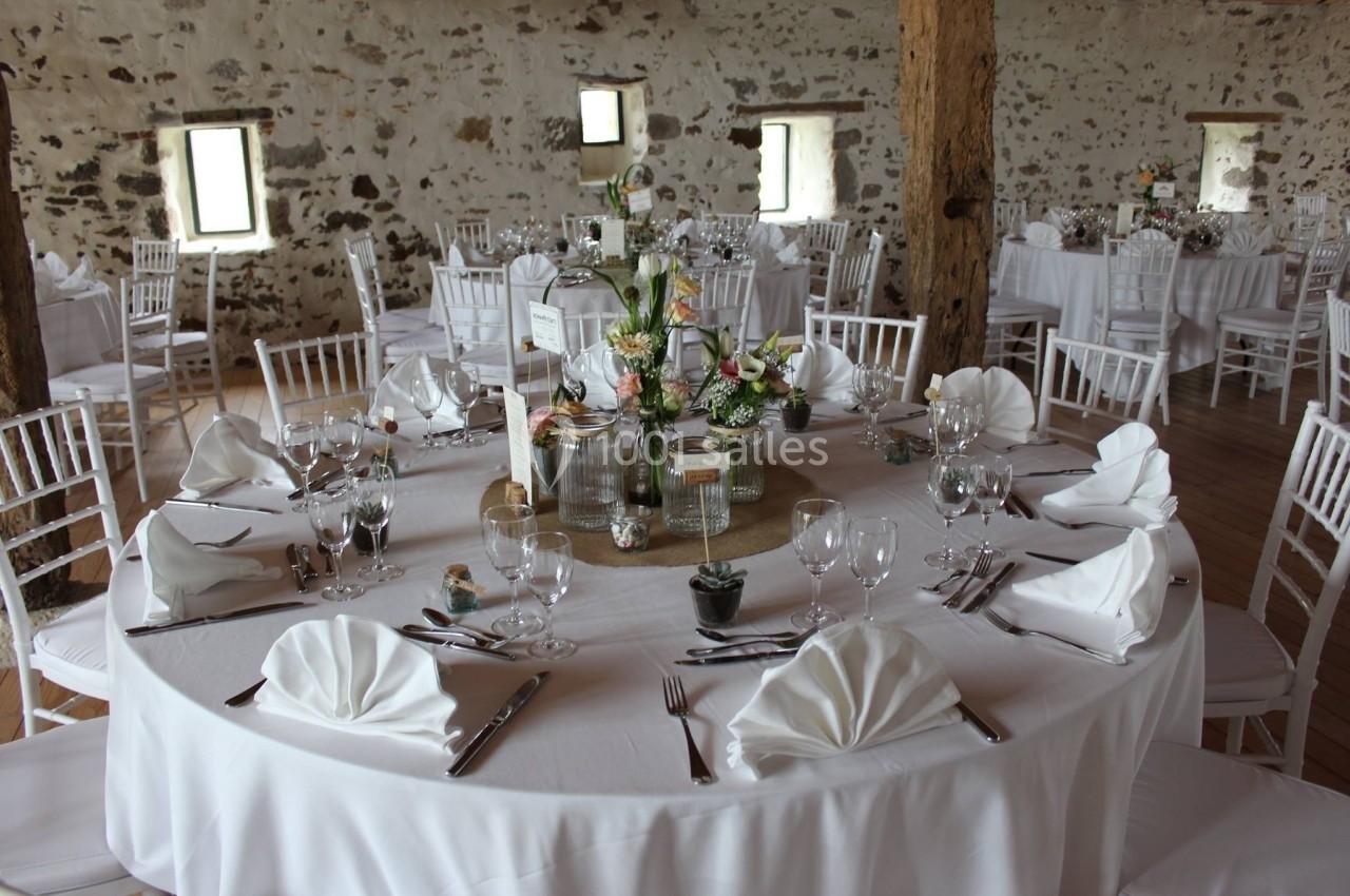 Salle de réception avec tables rondes dressées, nappes blanches, serviettes pliées et décoration florale centrale.