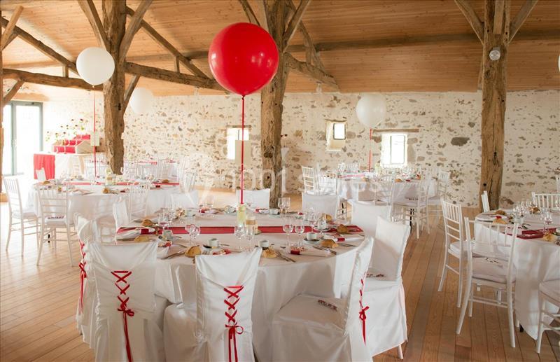 Salle de réception décorée avec des tables rondes, nappes blanches, chaises habillées et ballons rouges et blancs.