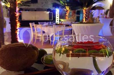 Espace salon lumineux avec fauteuils en osier, cheminée en pierre, piano, plantes et fenêtre donnant sur un jardin.