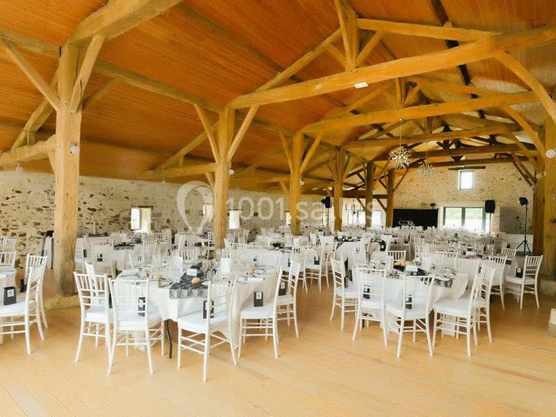 Salle de réception lumineuse avec poutres apparentes, tables rondes dressées et chaises blanches.