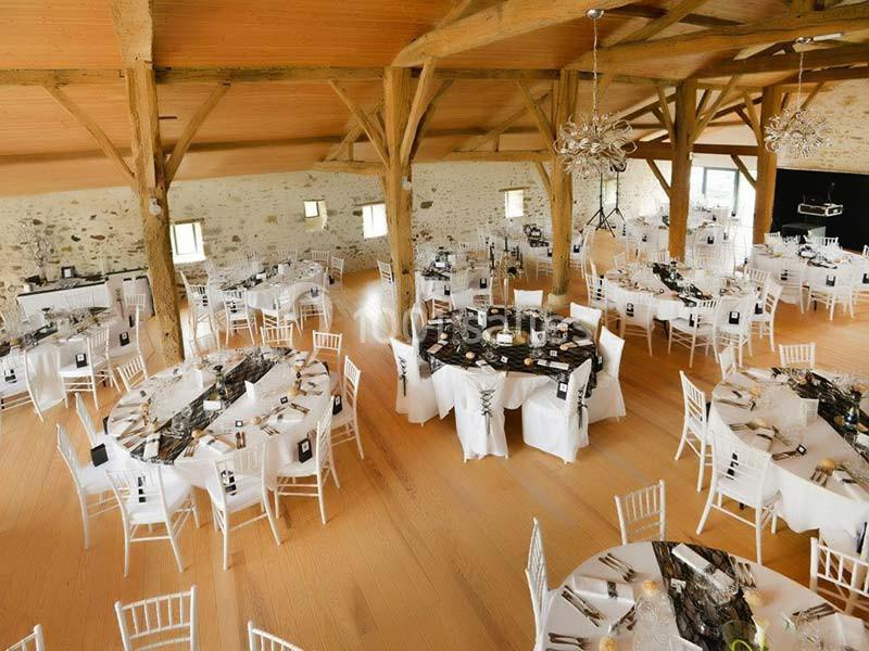 Salle de réception lumineuse avec tables rondes décorées, chaises blanches et poutres apparentes en bois.