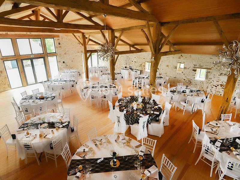 Salle de réception lumineuse avec poutres apparentes, tables rondes dressées et chaises blanches.
