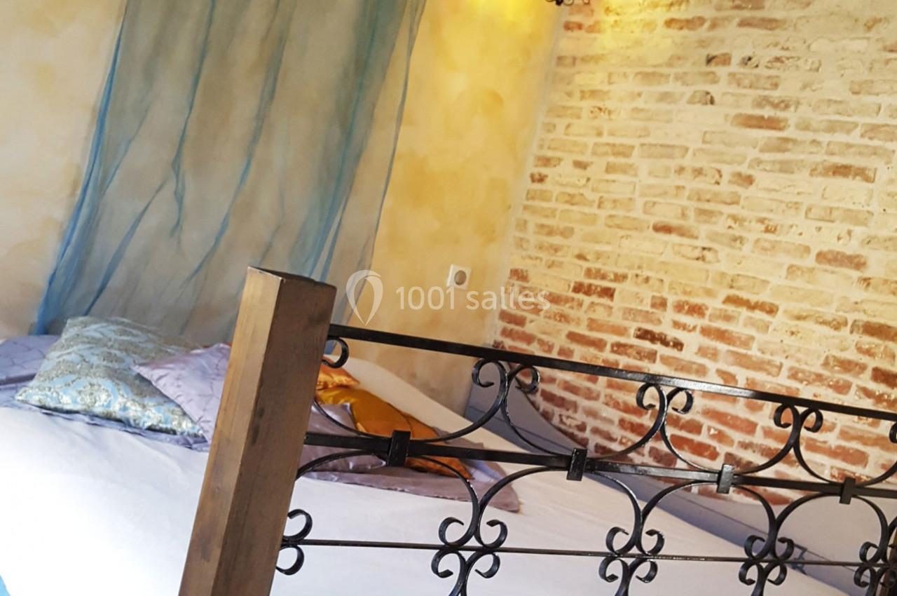 Lit double avec coussins colorés, tête de lit en fer forgé, mur en briques apparentes et rideau en voile bleu.