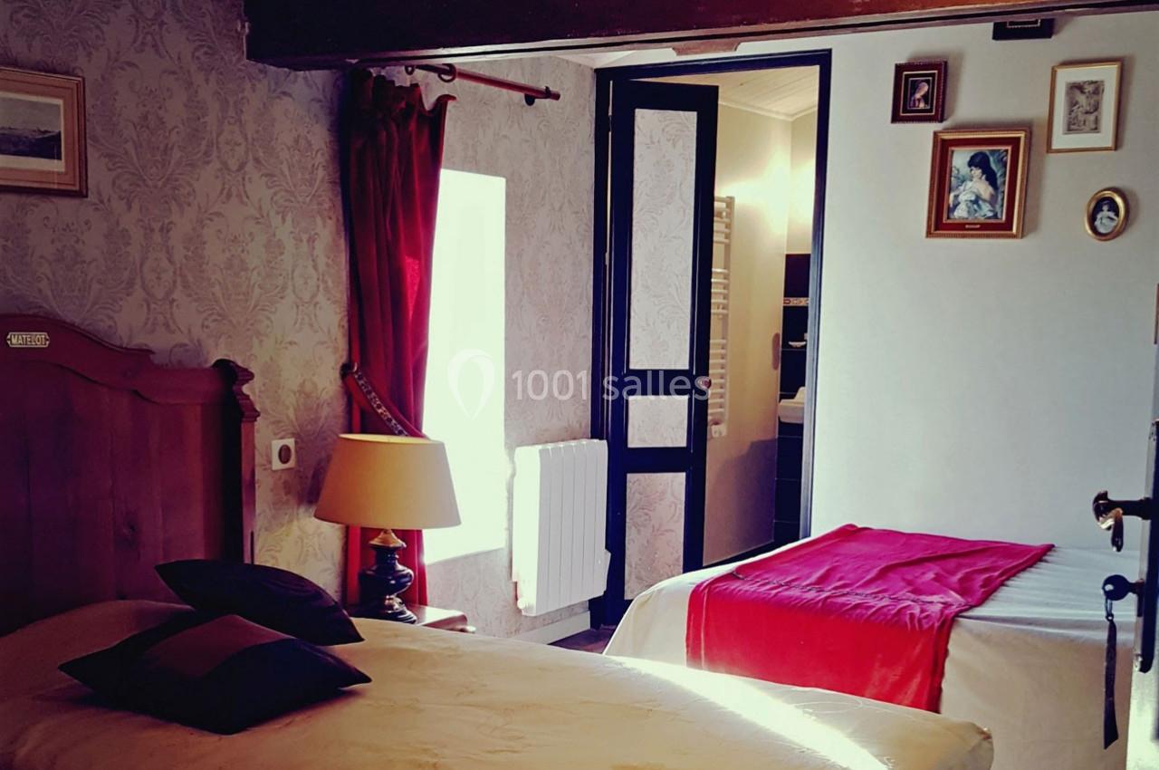 Chambre avec deux lits, murs décorés de cadres, rideaux rouges et lumière naturelle entrant par une fenêtre.