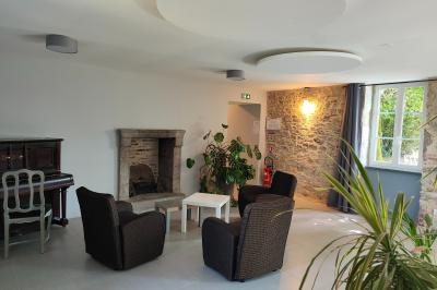 Espace salon lumineux avec fauteuils en osier, cheminée en pierre, piano, plantes et fenêtre donnant sur un jardin.