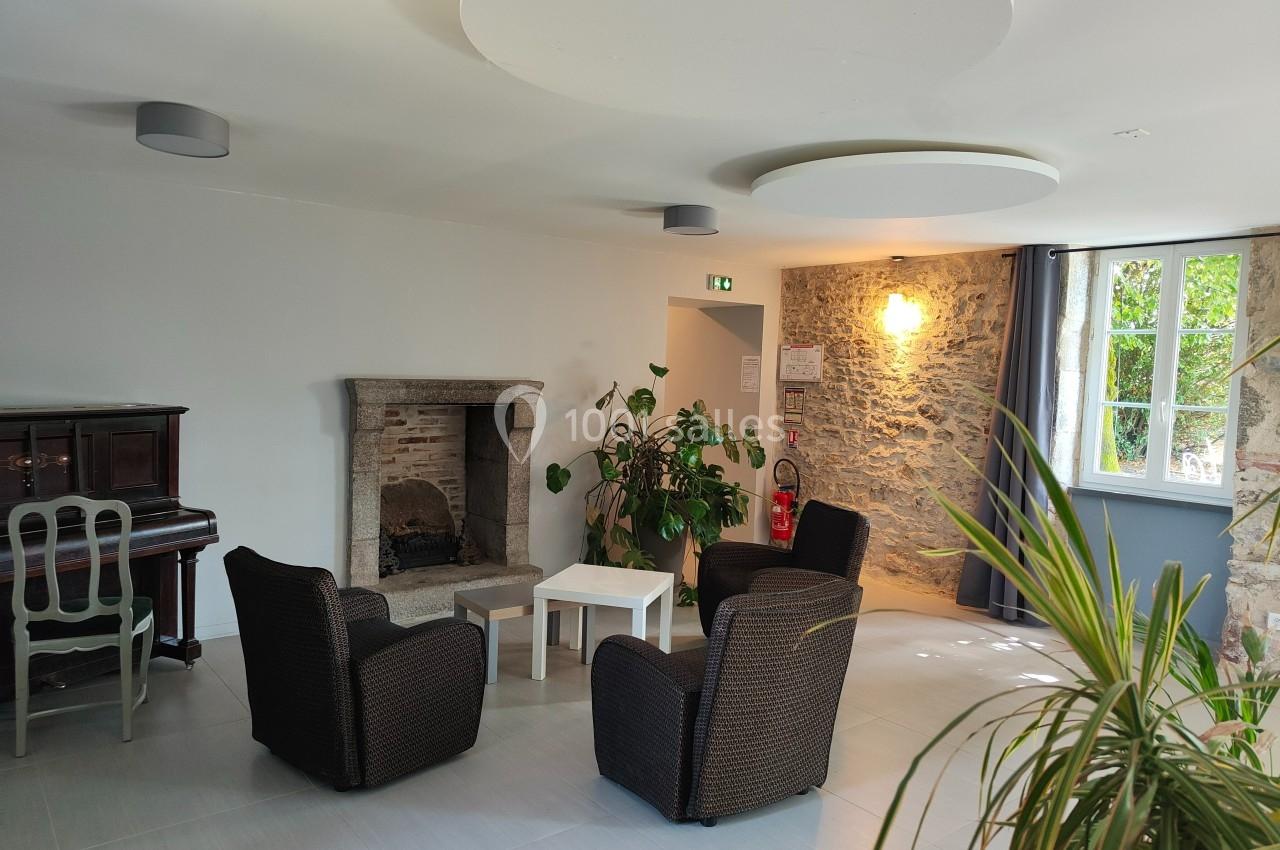 Espace salon lumineux avec fauteuils en osier, cheminée en pierre, piano, plantes et fenêtre donnant sur un jardin.