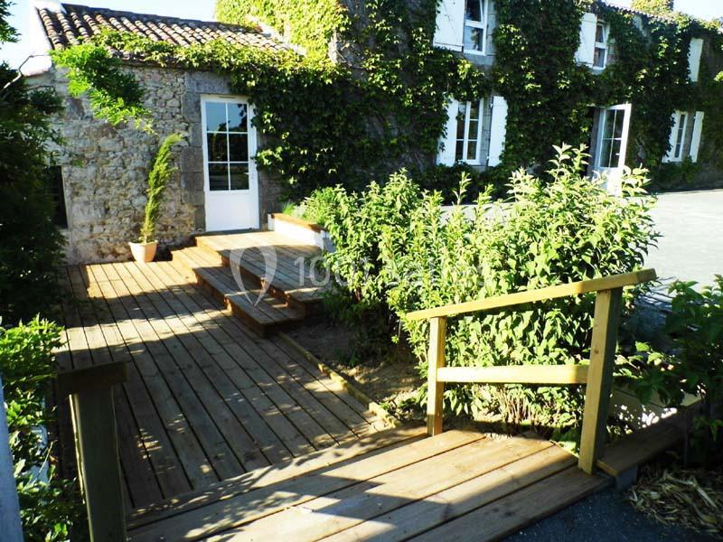 Passerelle en bois menant à une maison en pierre recouverte de lierre, avec terrasse et végétation environnante.