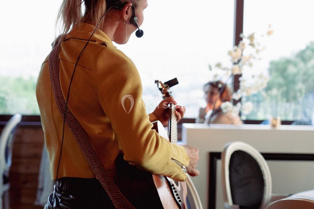 Une musicienne joue de la guitare avec un micro-casque dans un restaurant lumineux avec vue sur l'extérieur.