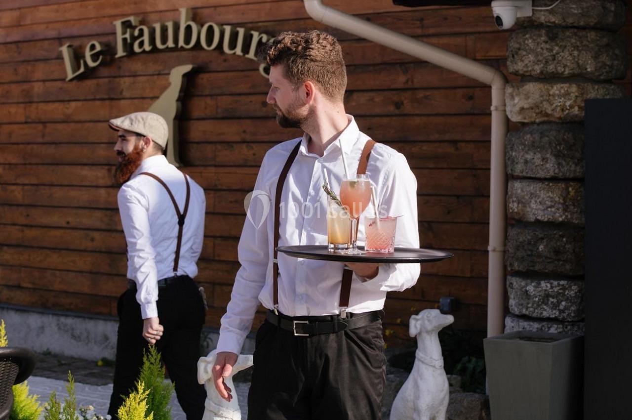 Un serveur en chemise blanche porte un plateau avec des boissons devant un bâtiment en bois nommé ’Le Faubourg’.