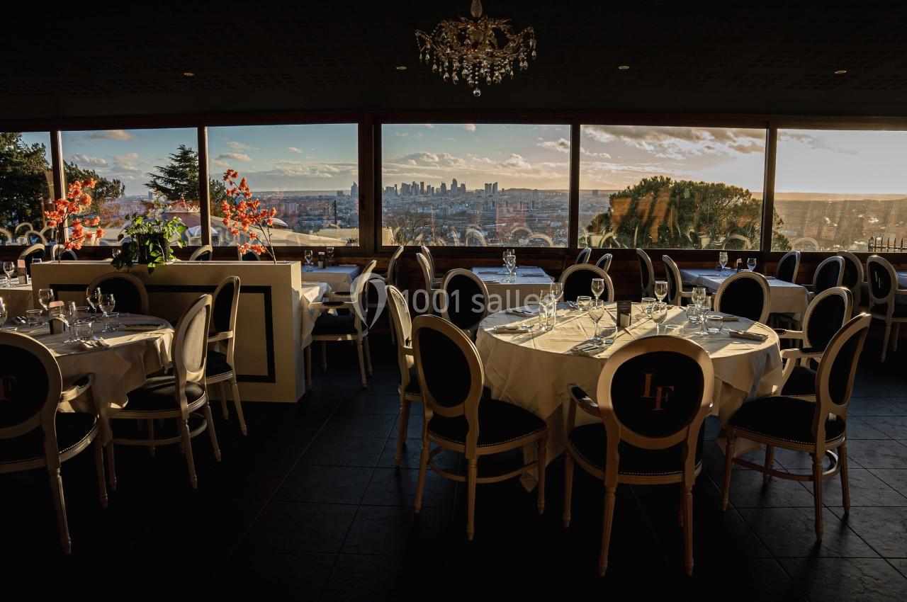 Salle de restaurant élégante avec des tables dressées, offrant une vue panoramique sur une ville au coucher du soleil.