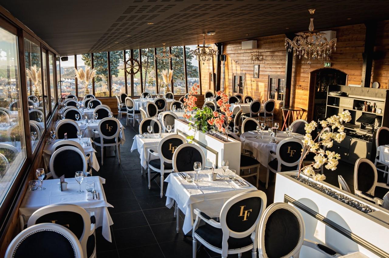 Salle de restaurant lumineuse avec de grandes baies vitrées, tables dressées et décoration élégante en bois et fleurs.