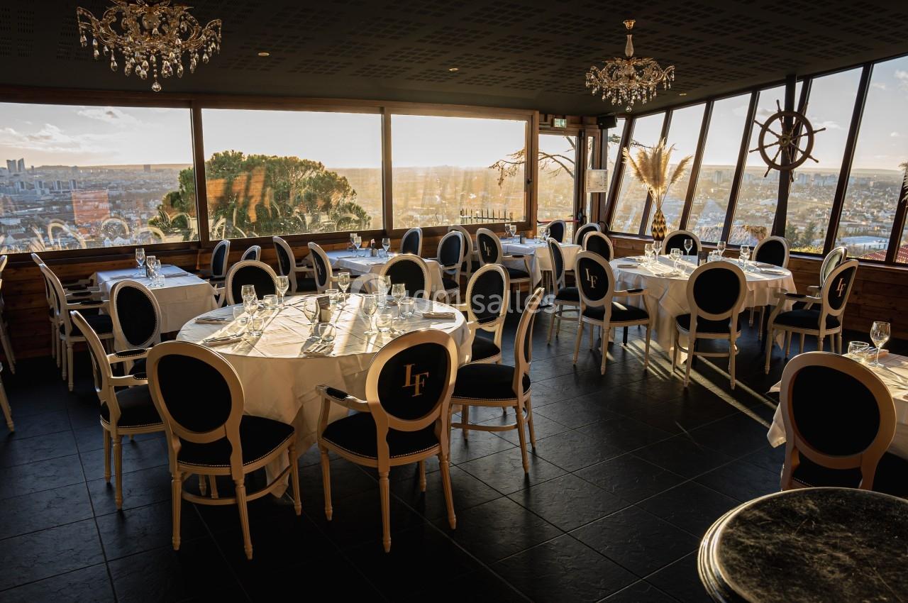 Salle de restaurant élégante avec grandes baies vitrées offrant une vue panoramique sur une ville au coucher du soleil.