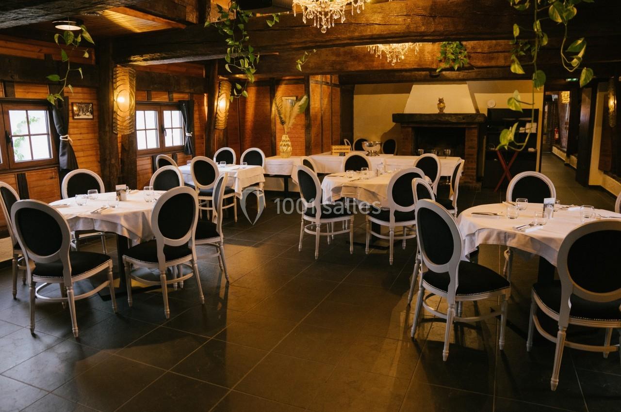Salle de restaurant chaleureuse avec tables rondes dressées, chaises noires et blanches, poutres apparentes et éclairage…