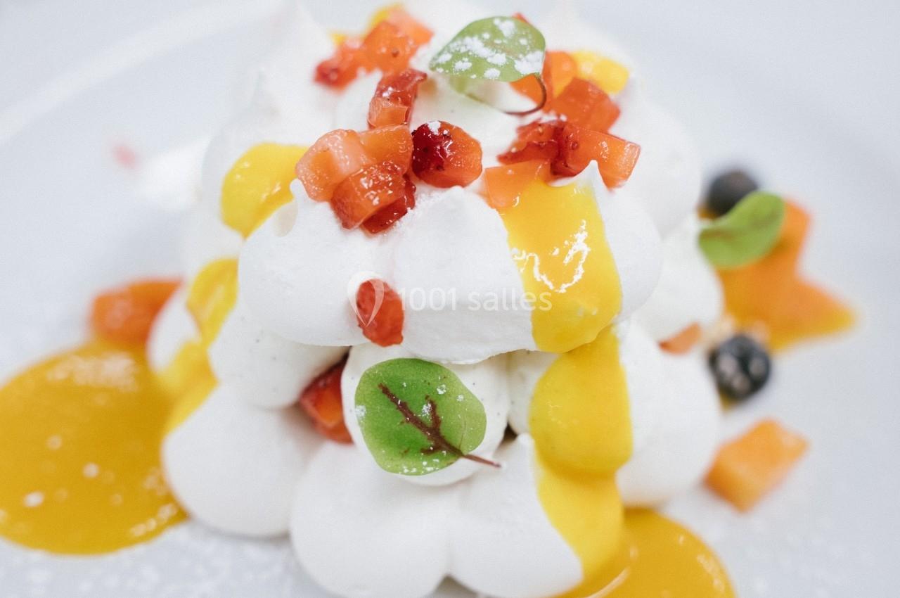 Dessert composé de meringue, coulis de mangue, morceaux de fruits rouges et feuilles décoratives sur une assiette blanche.