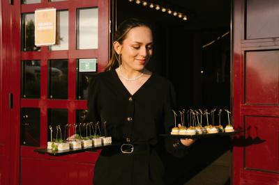 Une femme en tenue noire porte deux plateaux de hors-d'œuvre devant une porte rouge ouverte.