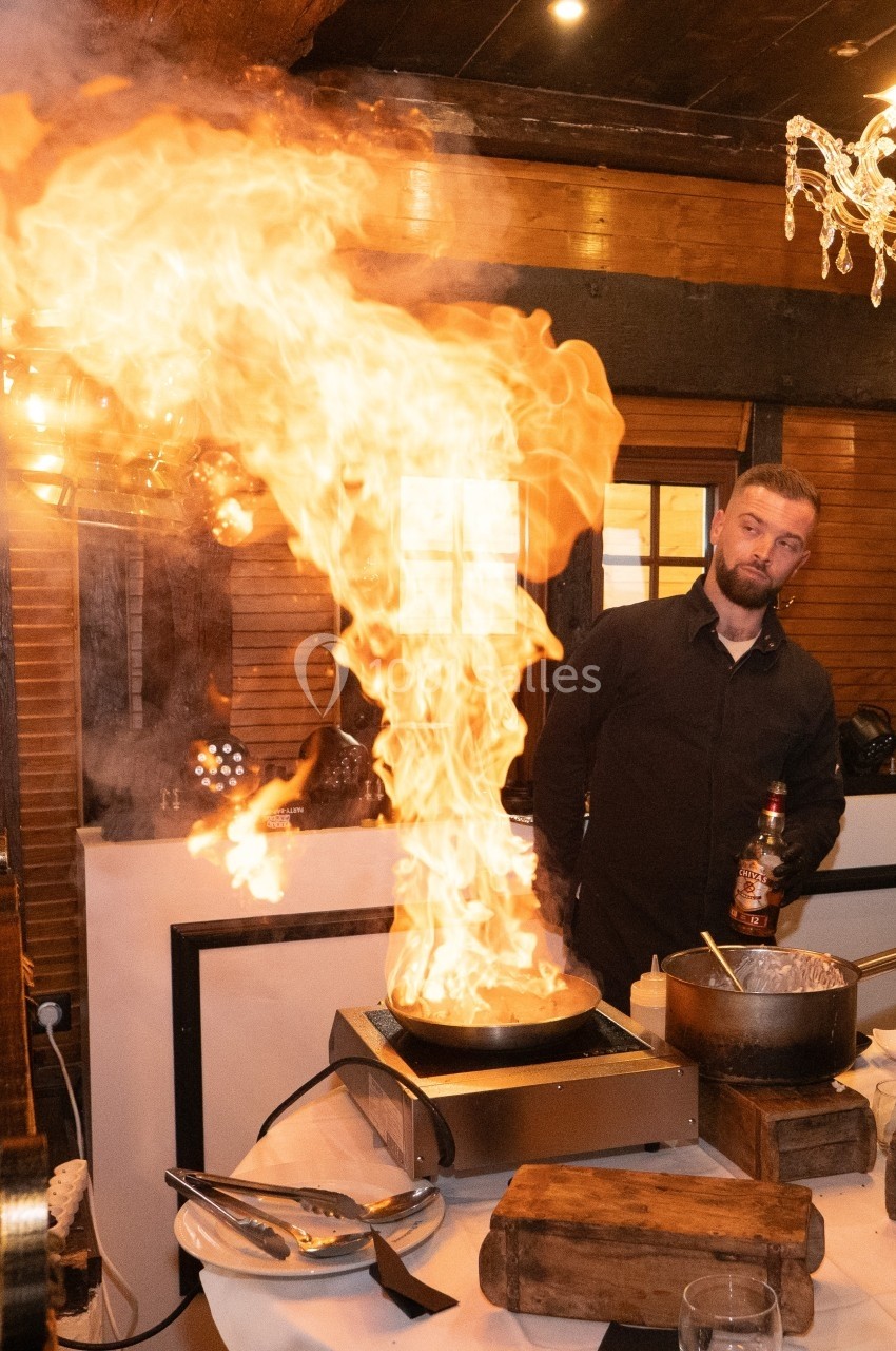 Un chef prépare un plat flambé avec une grande flamme dans une cuisine ouverte, sous un éclairage chaleureux.