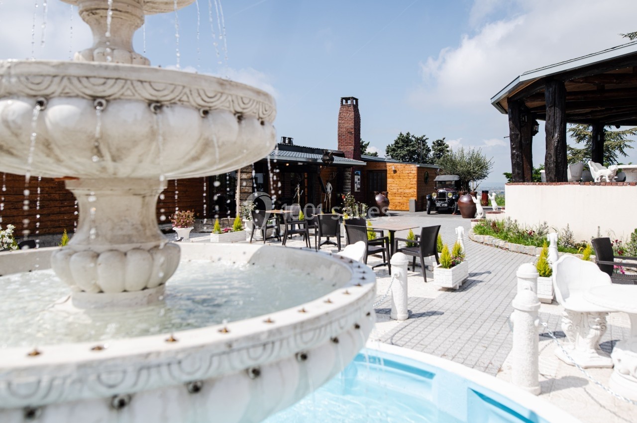 Fontaine blanche au premier plan dans une cour extérieure aménagée avec des tables, des chaises et des bâtiments en bois.