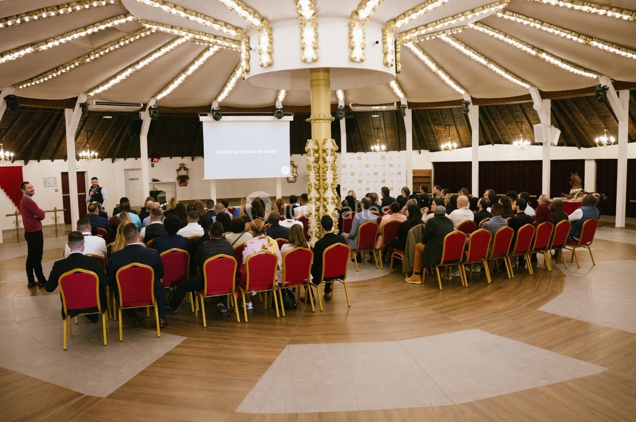 Salle de conférence circulaire avec des participants assis sur des chaises rouges, regardant une présentation projetée.