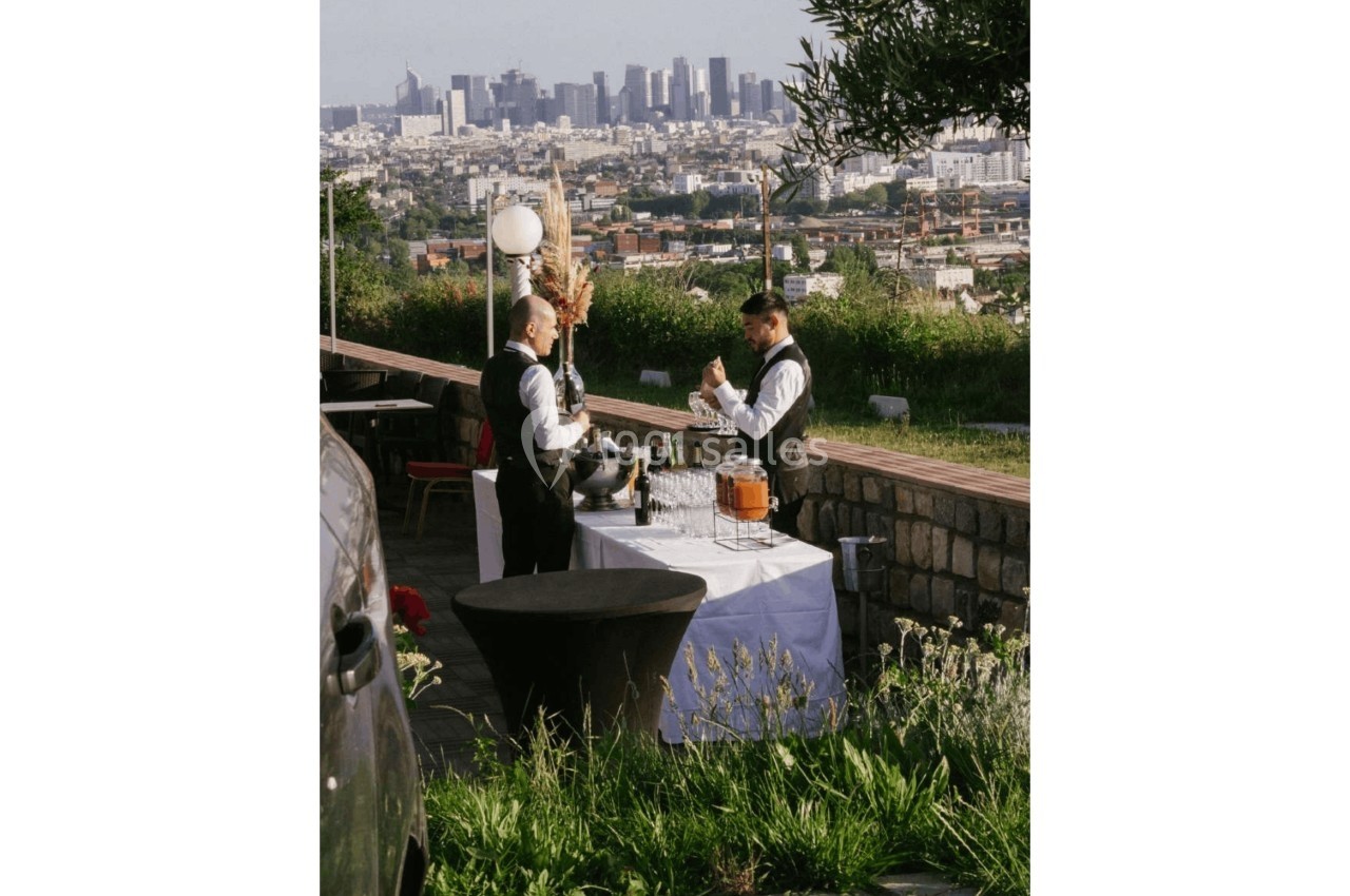 Deux serveurs en tenue formelle préparent des boissons à une table dressée en extérieur avec vue sur une ville.