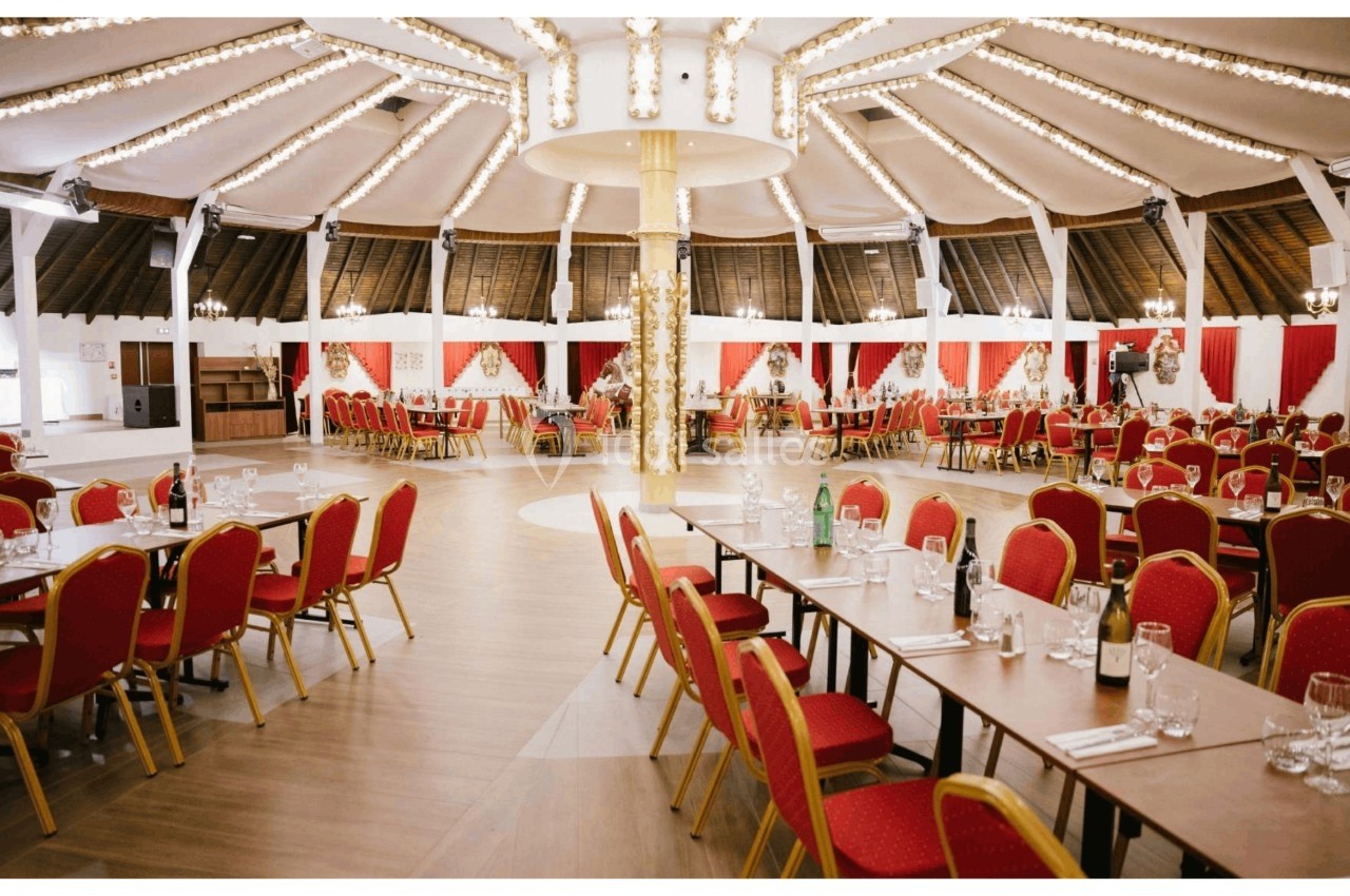 Salle de réception circulaire avec tables dressées, chaises rouges et éclairage suspendu sous un toit en bois.