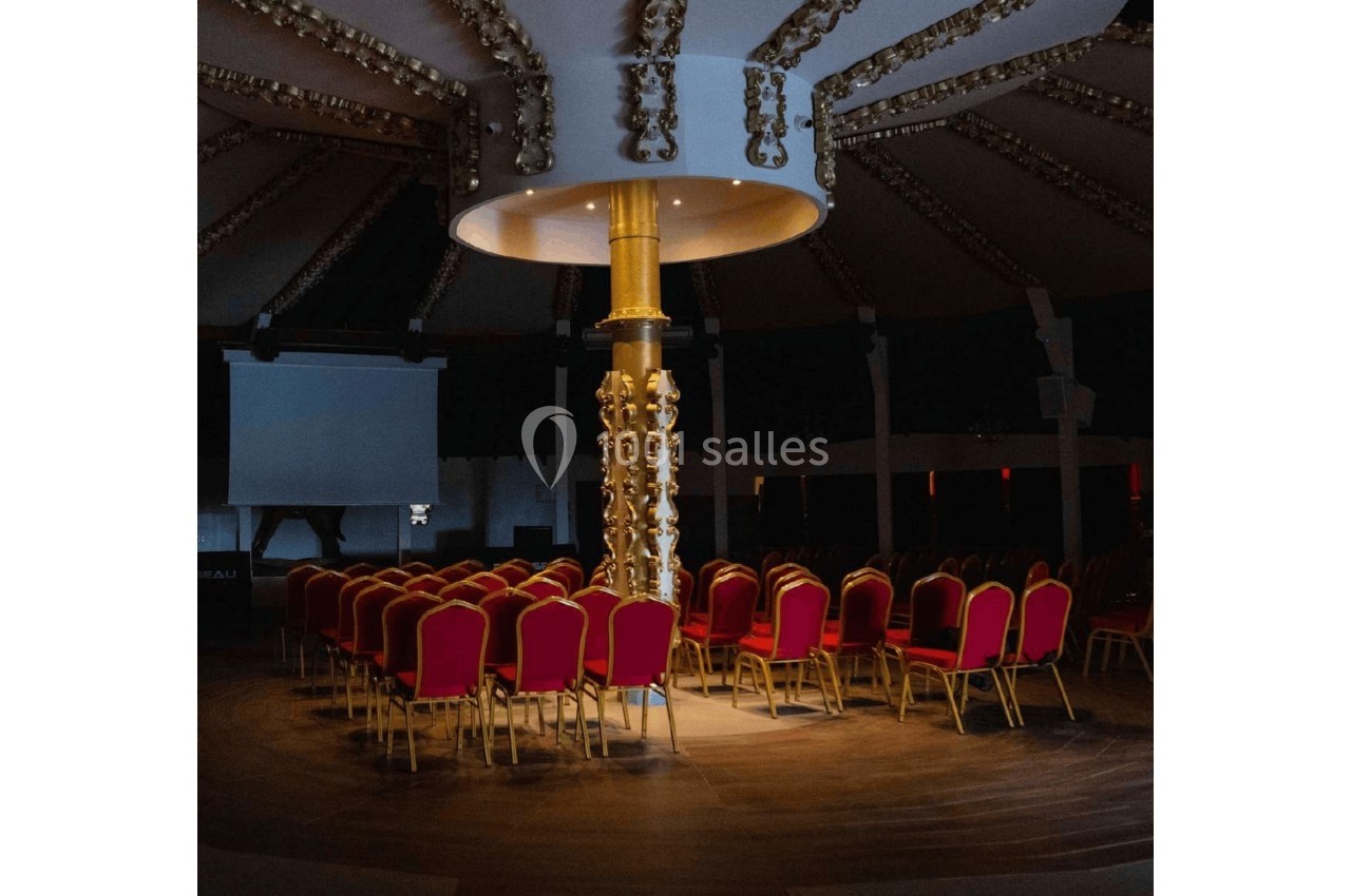 Chaises rouges disposées en cercle autour d'un pilier doré sous une lumière tamisée dans une salle sombre.