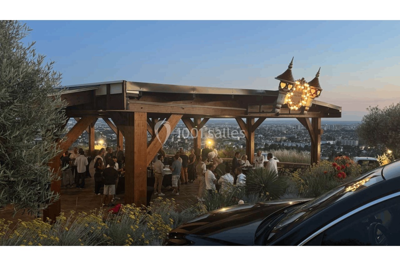 Terrasse en bois surélevée avec des convives, vue panoramique sur une ville au coucher du soleil.