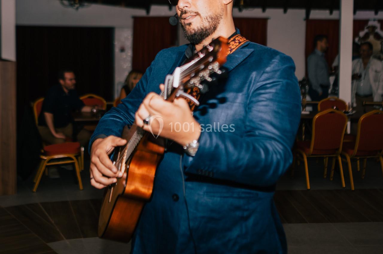Un homme en veste bleue joue de la guitare dans une salle avec des tables et des chaises rouges en arrière-plan.