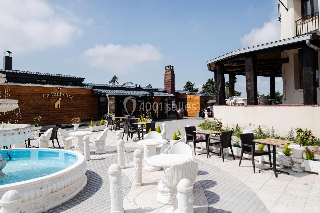 Terrasse extérieure avec tables, chaises, fontaine centrale et bâtiments en arrière-plan sous un ciel dégagé.