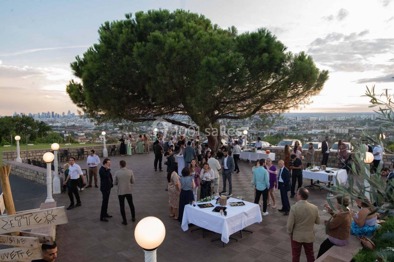 Groupe de personnes rassemblées sur une terrasse avec vue panoramique, autour de tables, sous un grand pin au coucher du…