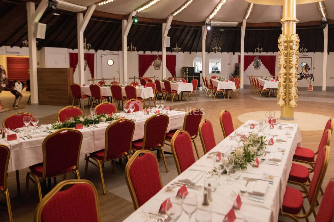 Salle de réception décorée avec des tables dressées, nappes blanches, chaises rouges et ornements végétaux.