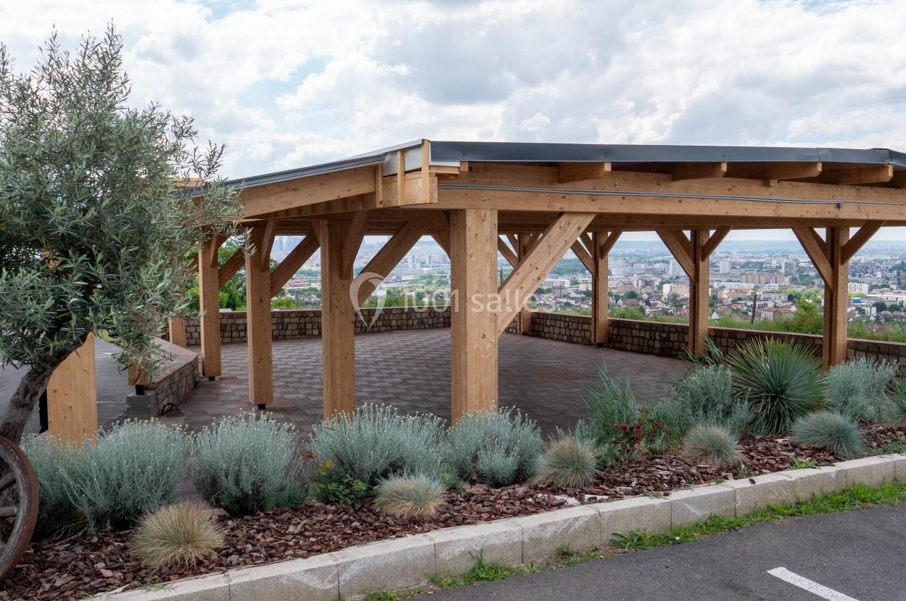 Structure en bois ouverte avec toit, entourée de plantes et offrant une vue panoramique sur une ville en arrière-plan.