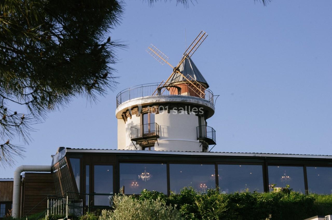 Un moulin restauré avec des ailes en bois surmontant un bâtiment moderne entouré de végétation.