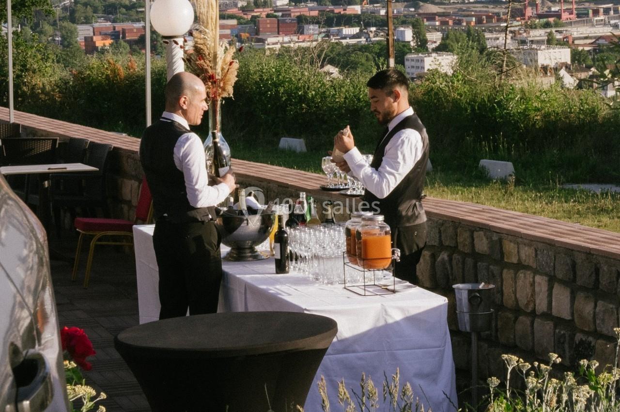 Deux serveurs en tenue formelle préparent une table avec boissons et verres, en extérieur avec vue sur une ville.