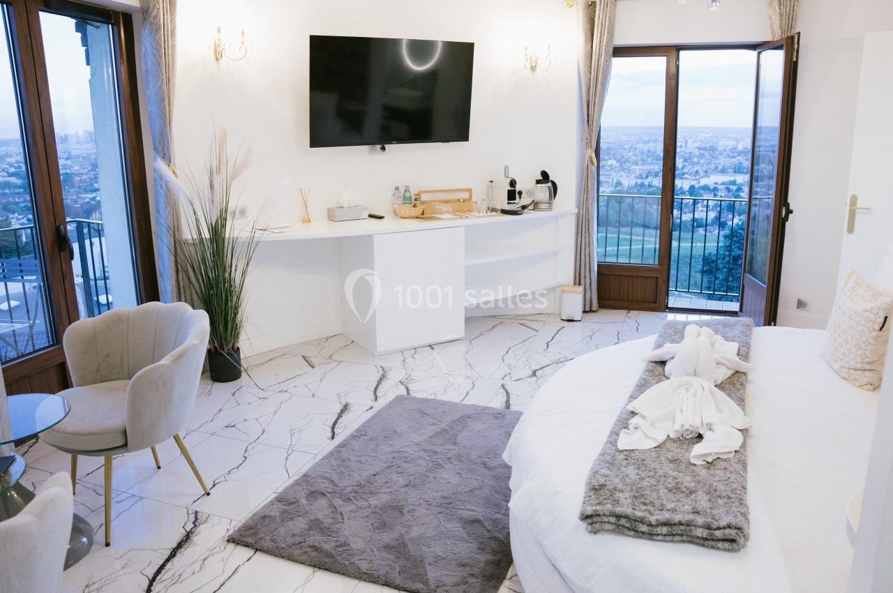 Chambre lumineuse avec lit double, télévision murale, fauteuils, et vue panoramique sur une ville depuis un balcon.