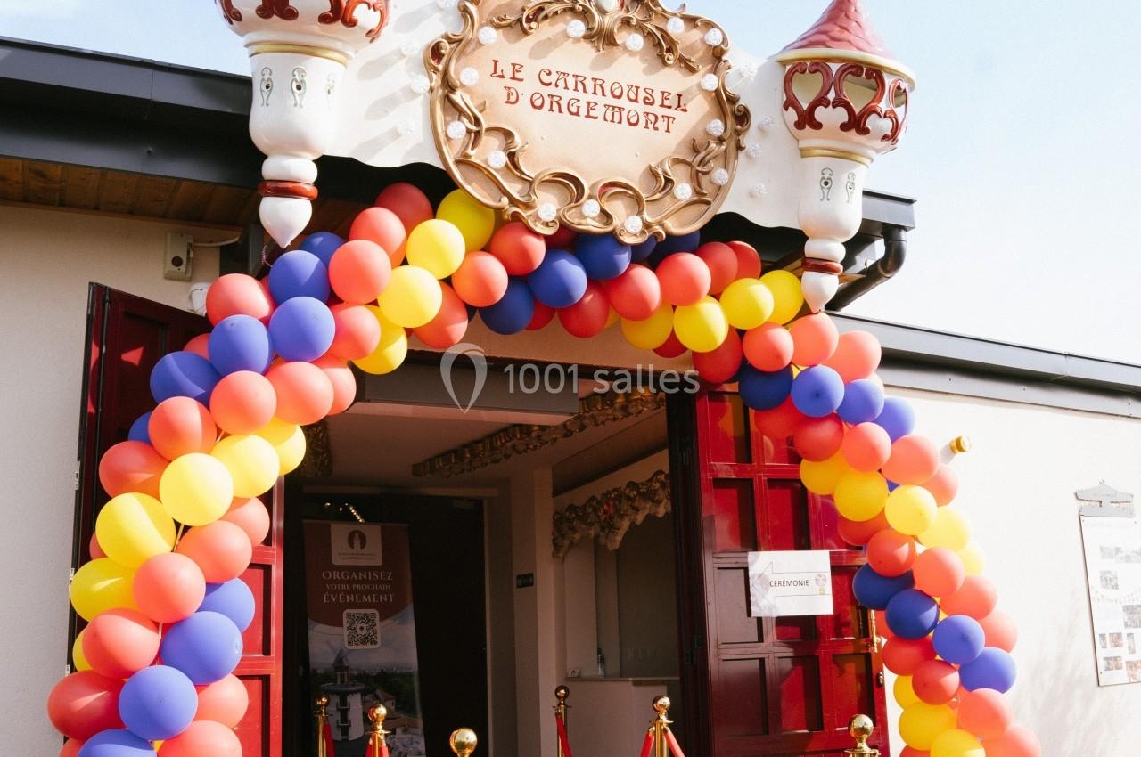 Entrée décorée avec une arche de ballons colorés et un tapis rouge menant à un bâtiment au style festif.
