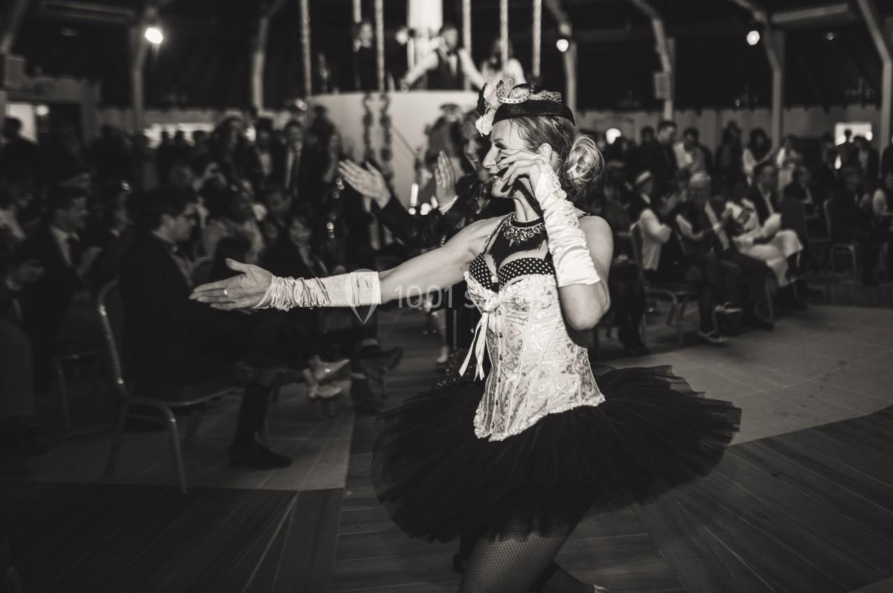 Danseuse en tenue de cabaret tenant un micro, performe devant un public assis dans une salle sombre.