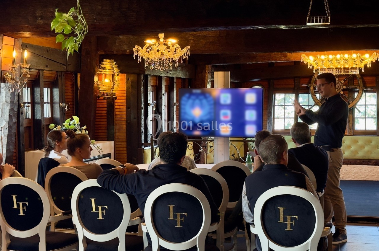Un homme présente un écran interactif à un groupe de personnes assises dans une salle au décor rustique et élégant.
