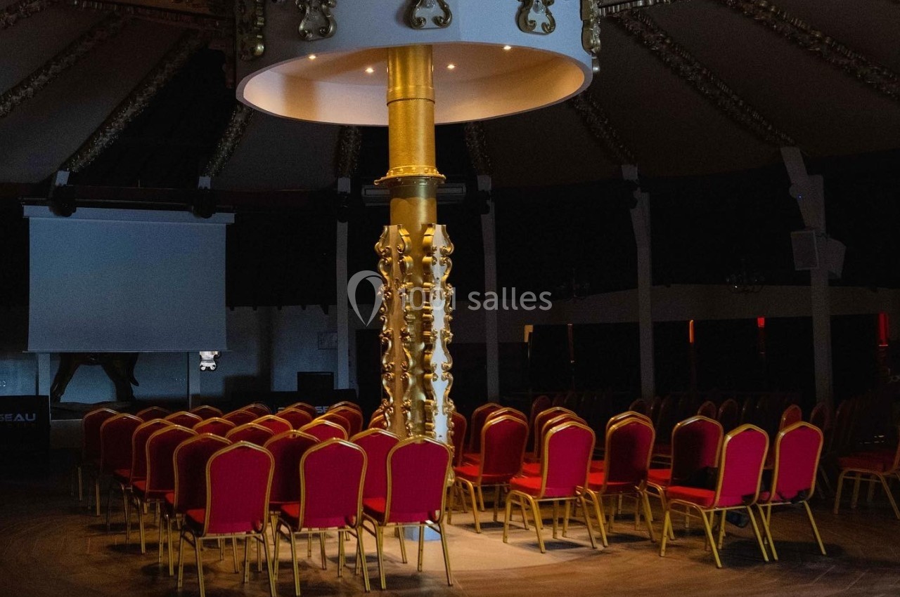 Chaises rouges disposées en cercle autour d'une colonne dorée dans une salle sombre avec un éclairage central.
