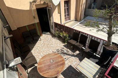 Terrasse ensoleillée avec table ronde en bois, chaises en rotin, plantes et voiles d'ombrage dans un cadre urbain.