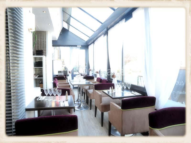 Salle lumineuse d'un restaurant avec tables dressées, chaises rembourrées et grandes baies vitrées laissant entrer la…