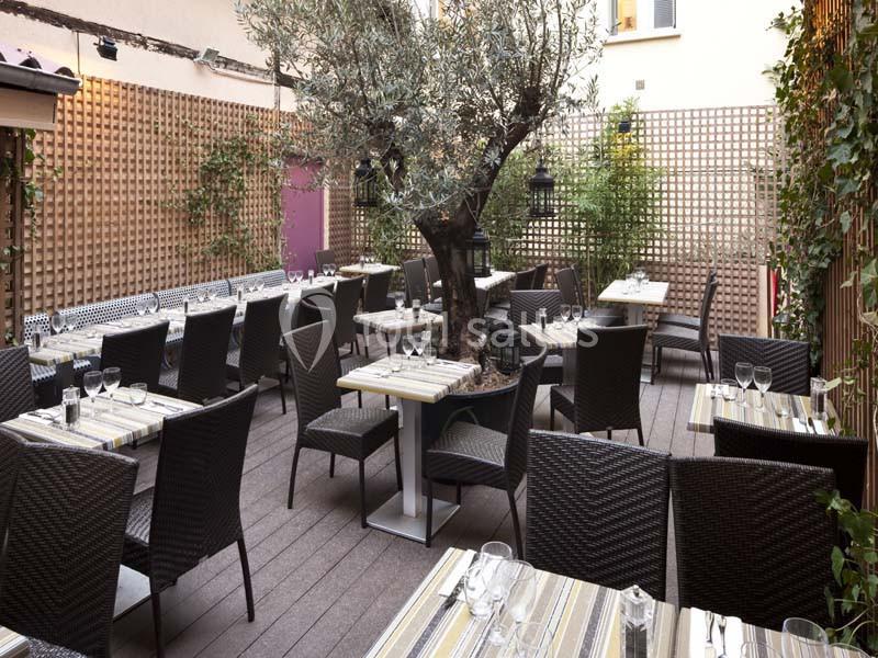 Terrasse extérieure avec tables dressées, chaises en osier, un olivier central et des panneaux en bois ajourés.