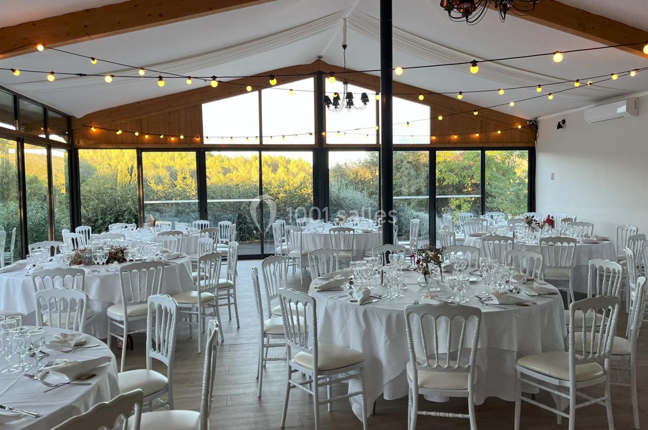 Salle de réception lumineuse avec tables rondes dressées, chaises blanches et guirlandes lumineuses suspendues.