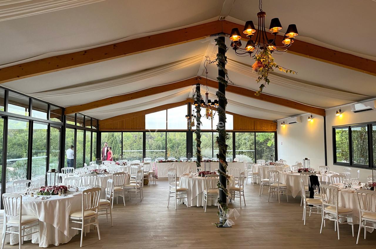 Salle de réception lumineuse avec tables rondes dressées, chaises blanches et décoration florale suspendue.
