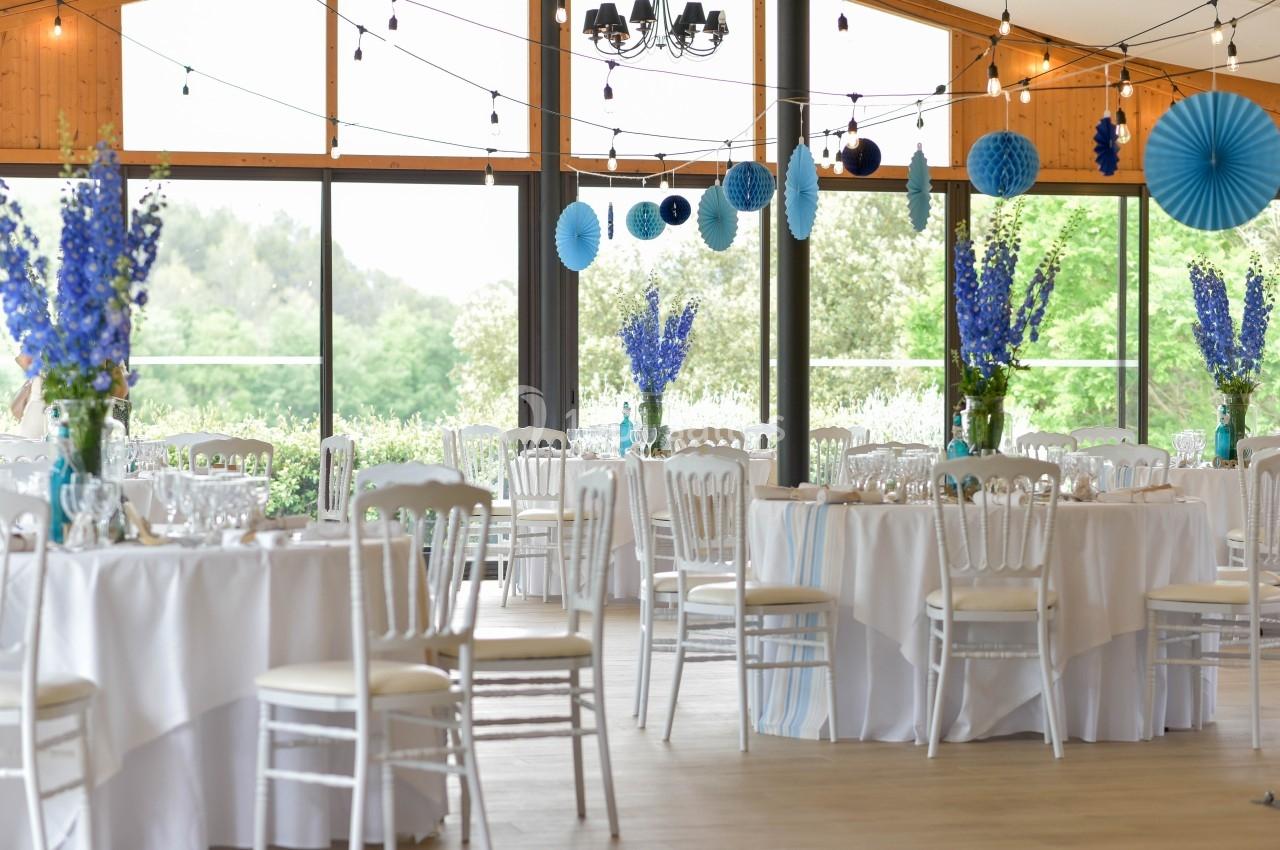 Salle de réception lumineuse décorée de guirlandes, fleurs bleues et tables rondes dressées avec nappes blanches.