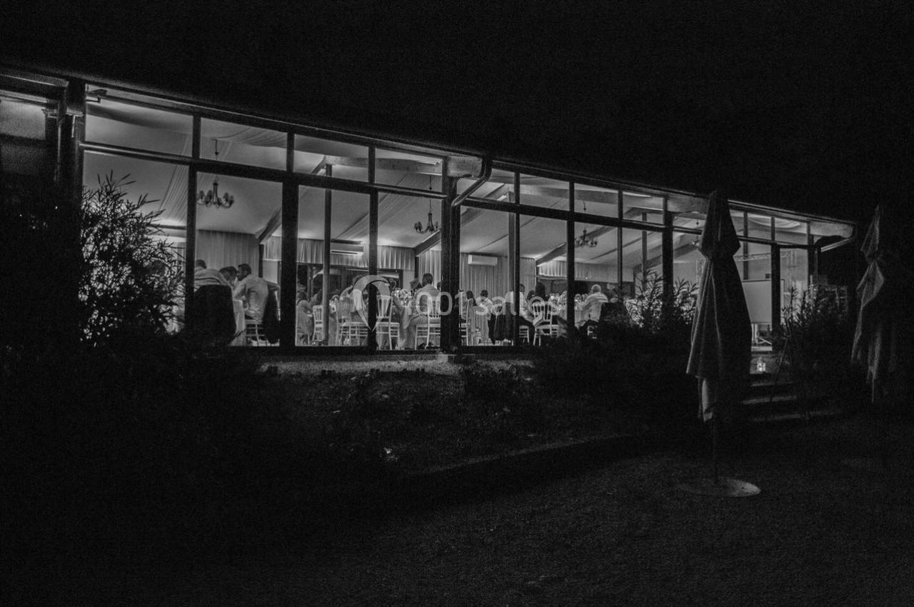 Vue nocturne d'une salle vitrée éclairée où des personnes sont réunies autour de tables.