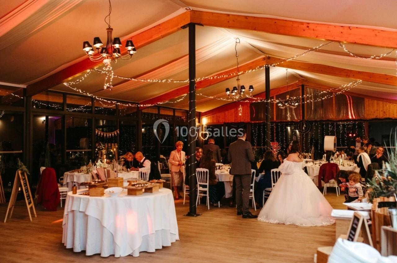 Salle de réception décorée avec des guirlandes lumineuses, des invités assis et debout, et une mariée en robe blanche.