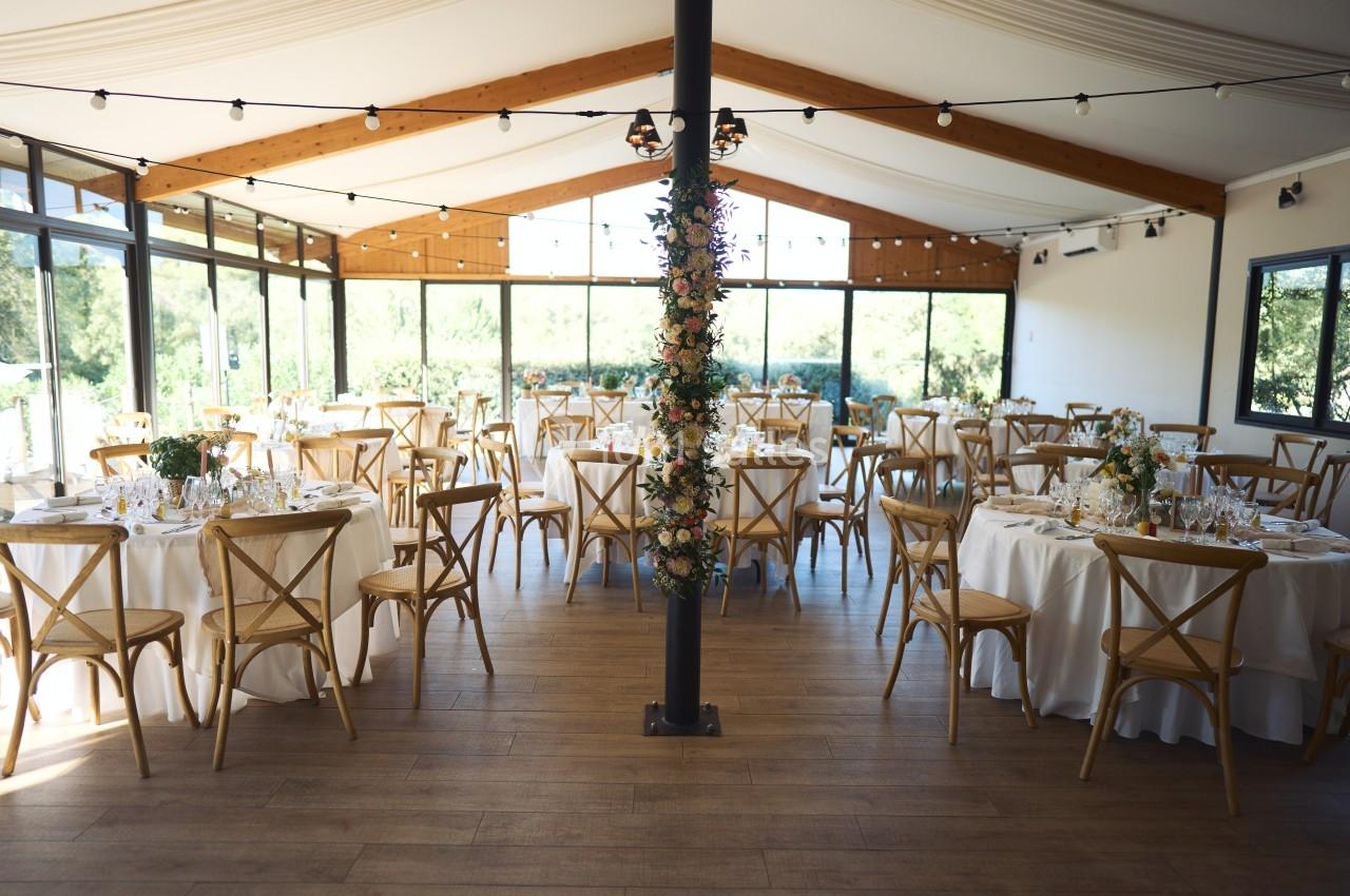 Salle de réception lumineuse avec tables rondes dressées, chaises en bois et décoration florale sur une colonne centrale.